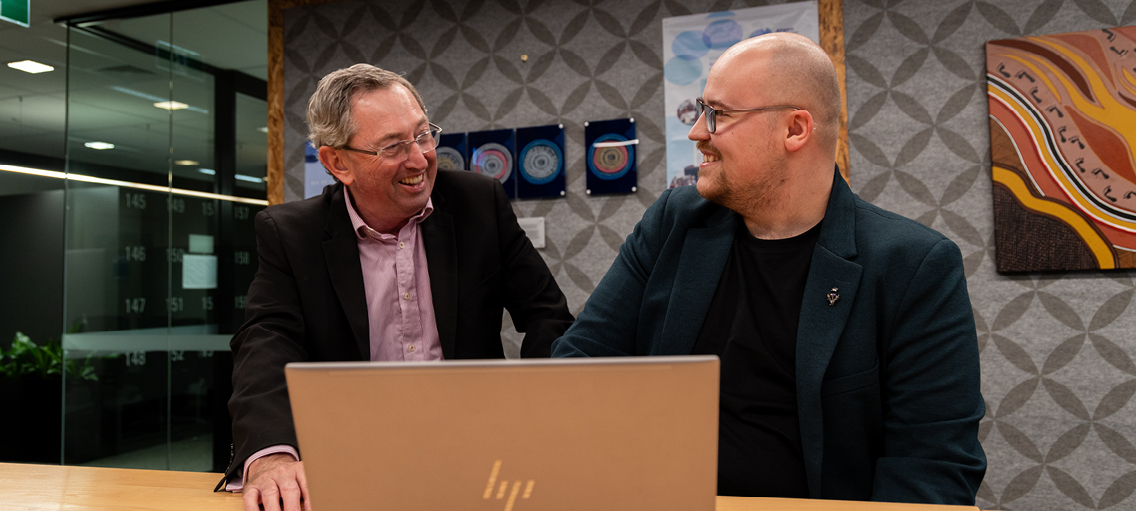 Photo of two staff member sitting at their desk smiling while reviewing work on a laptop